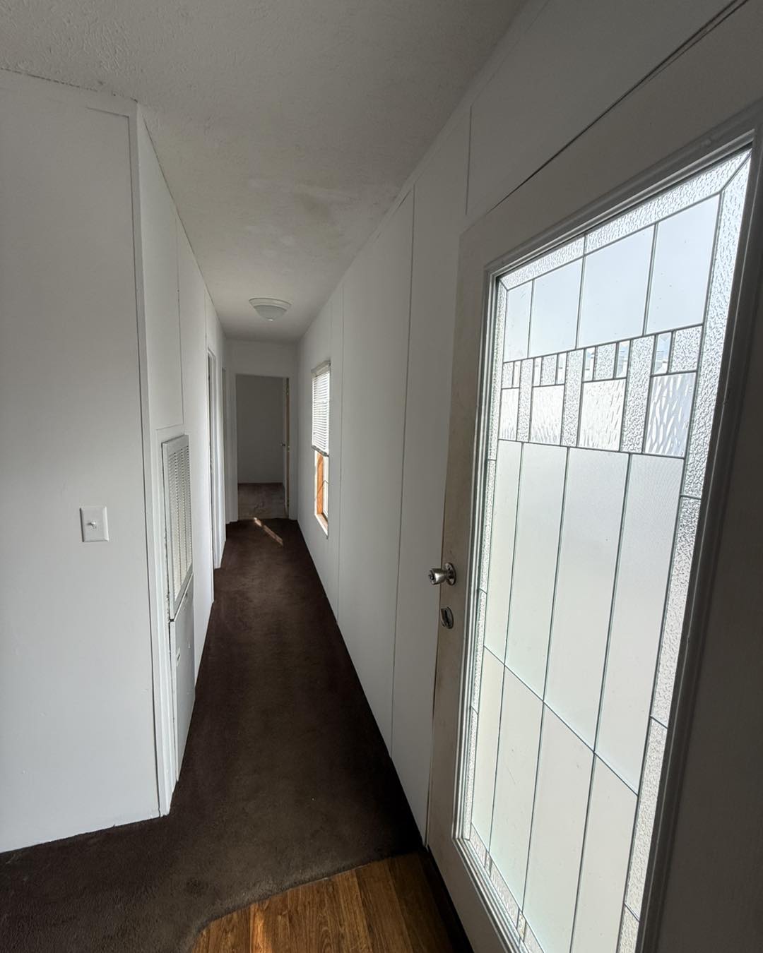 A narrow, dimly lit hallway with white walls and a brown carpet features frosted glass on the right door, a distant open door, and a broom on the floor.