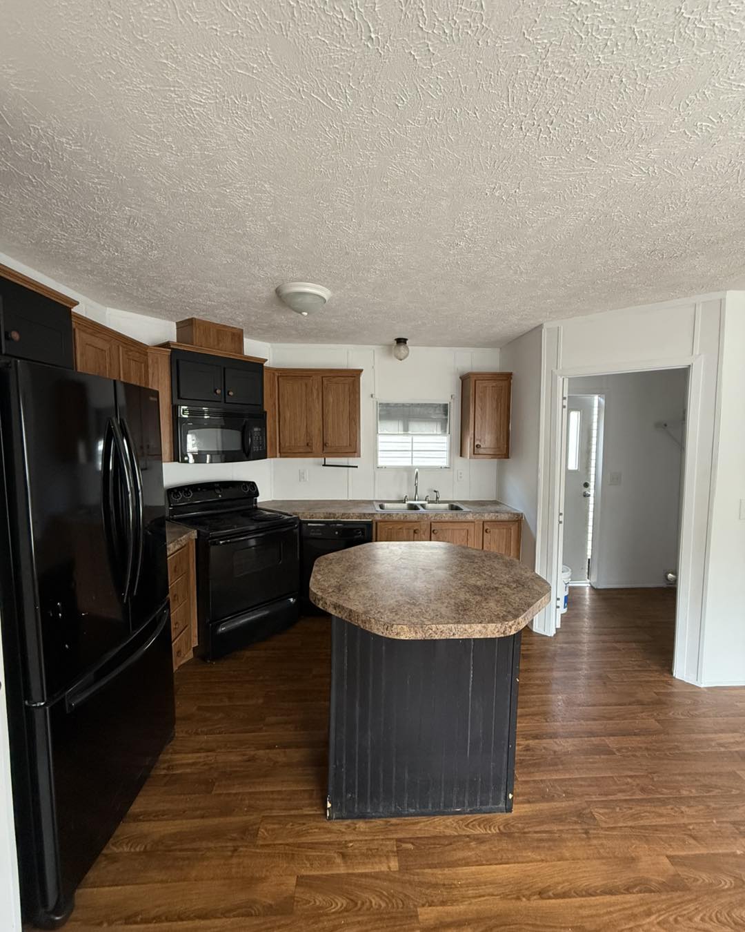 A cozy kitchen features wood cabinets, black appliances, and a central island with a stone countertop. The room has a warm, inviting atmosphere.