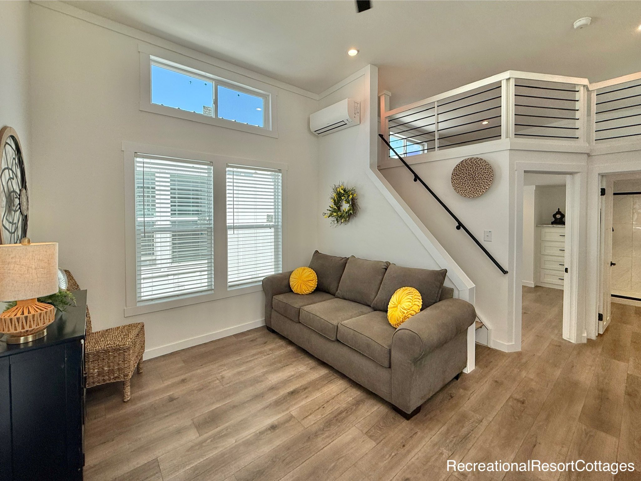 Modern living room with a gray sofa adorned with yellow pillows, wooden flooring, and bright windows. A staircase leads to an upstairs loft. Cozy and inviting atmosphere.