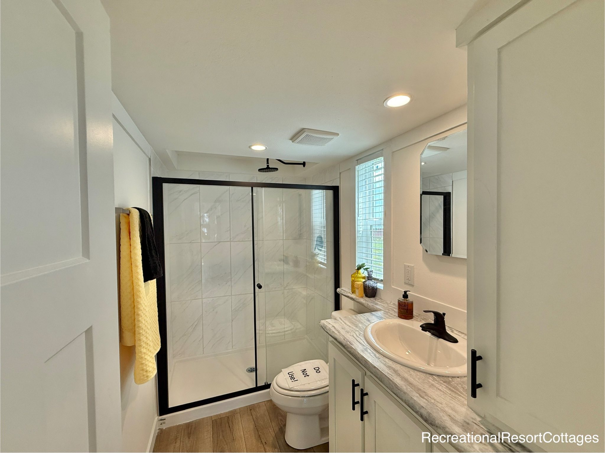 Modern bathroom featuring a glass shower with black trim, a white sink with a marble countertop, bright lighting, and yellow towels. Cozy and clean ambiance.