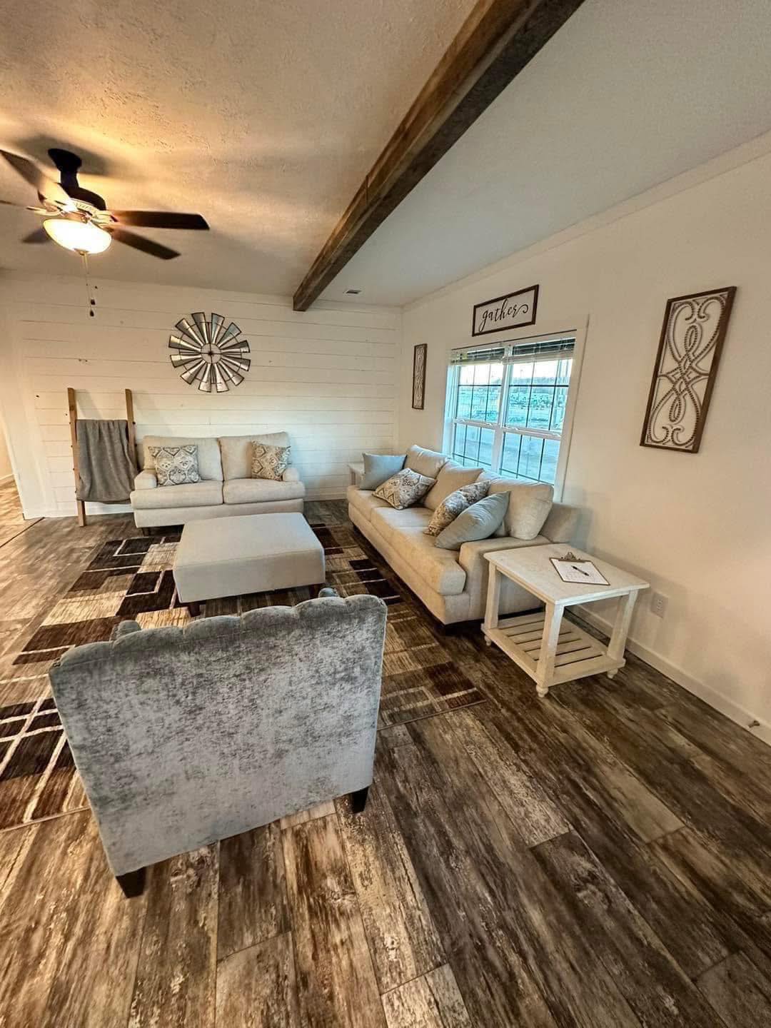 Cozy living room with cream sofas, patterned cushions, and a geometric rug. A ceiling fan spins above. Bright window light and rustic decor enhance warmth.