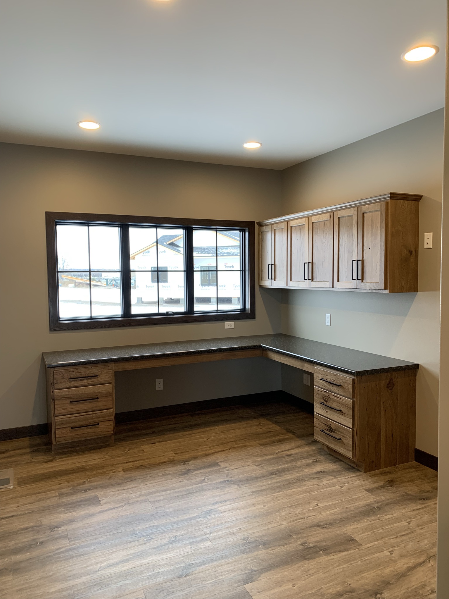 Corner office space with L-shaped wooden desk, matching overhead cabinets, and large window. Warm lighting and wood-patterned flooring create a cozy feel.