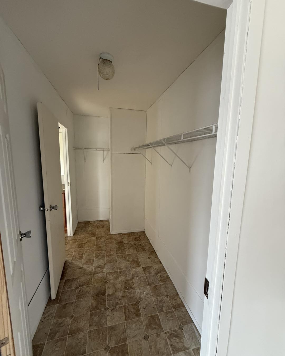 Empty narrow white closet with tiled floor, open door, and wire shelves along both walls, creating a stark, minimalist atmosphere.