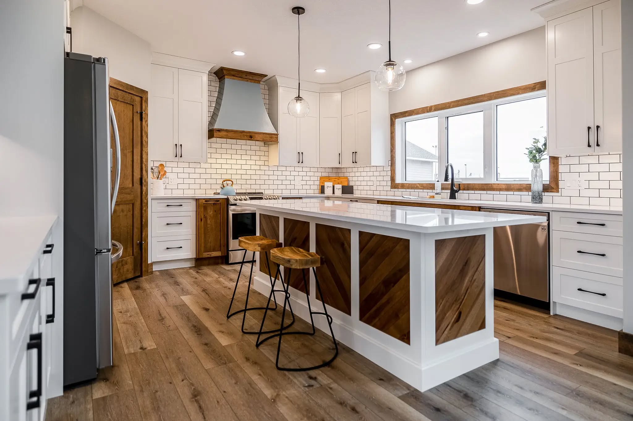 Modern kitchen with a large island featuring wood accents and white countertops. Pendant lights hang above, and bar stools sit in front. Bright and airy with subway tile backsplash, wooden floor, and stainless steel appliances.