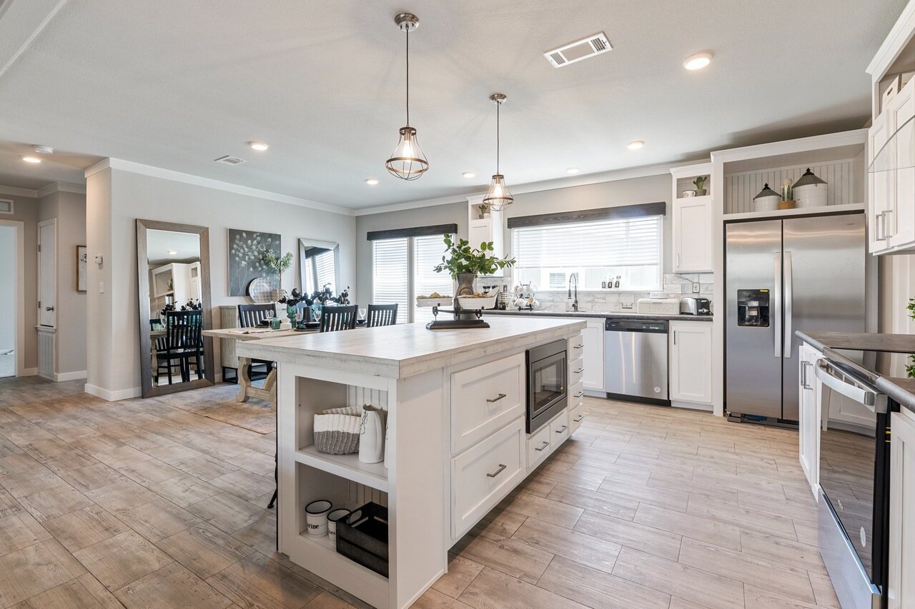 Bright, modern kitchen with a central island, pendant lights, stainless steel appliances, and wooden flooring. Open dining area is visible in the background.