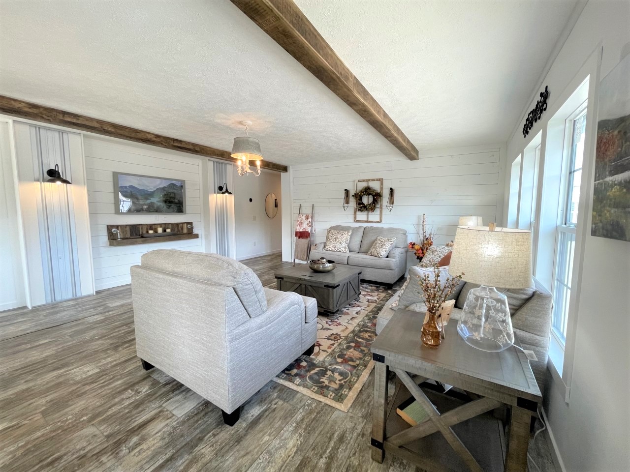Spacious living room with light wood floors, gray sofas, rustic wood beam ceiling, and large windows. Decor includes a wreath and warm lighting.