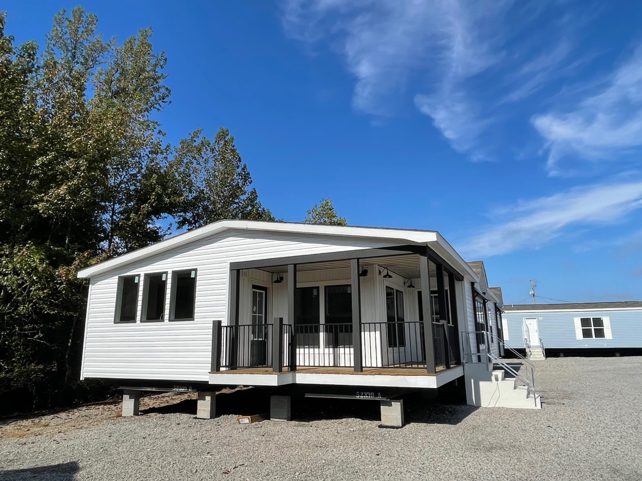 White mobile home with a front porch and black railings, set on a gravel lot under a clear blue sky. Trees are visible in the background.