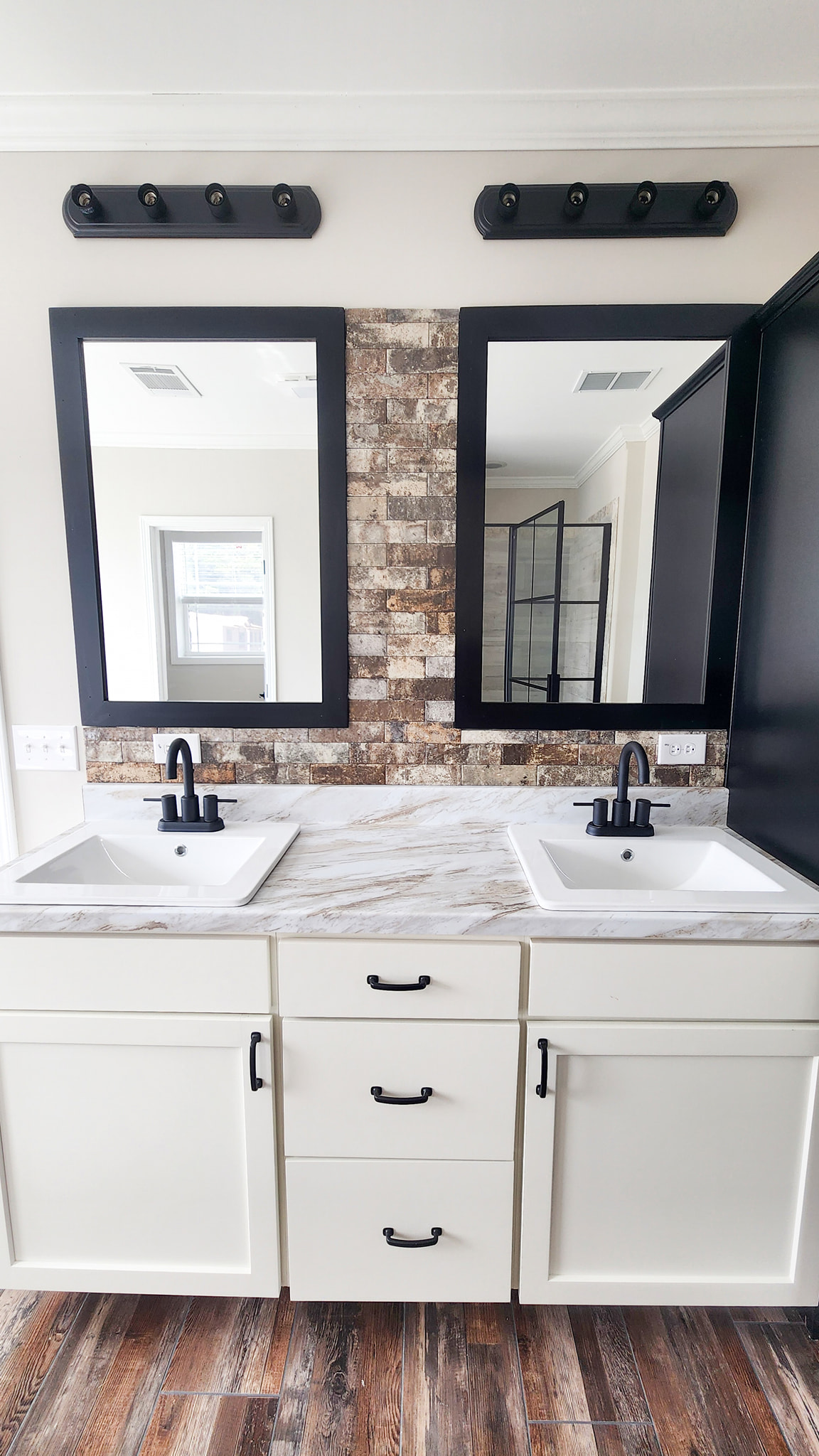 Modern bathroom with dual sinks on a white marble countertop. Two black-framed mirrors hang above, with rustic brick backsplash and black faucets, creating a sleek, contemporary feel.