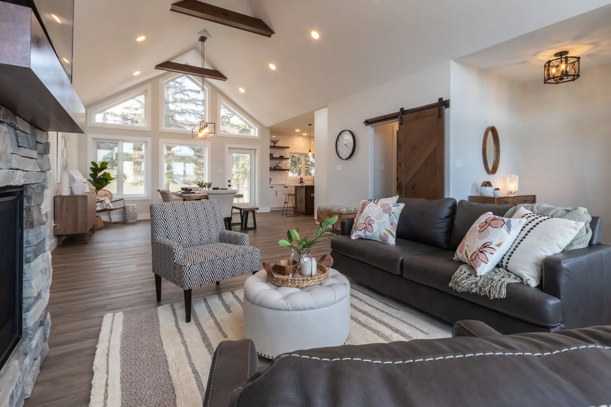 A modern living room with high ceilings, exposed beams, and large windows. Features a dark leather sofa, patterned armchair, and stone fireplace, creating an inviting, cozy ambiance.