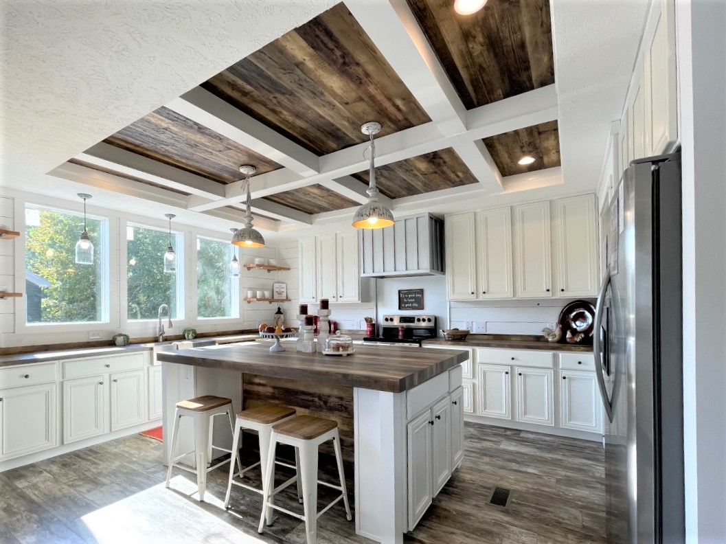 Spacious farmhouse kitchen with white cabinets and rustic wood accents. Features an island with stools, hanging lights, and large windows with greenery outside.