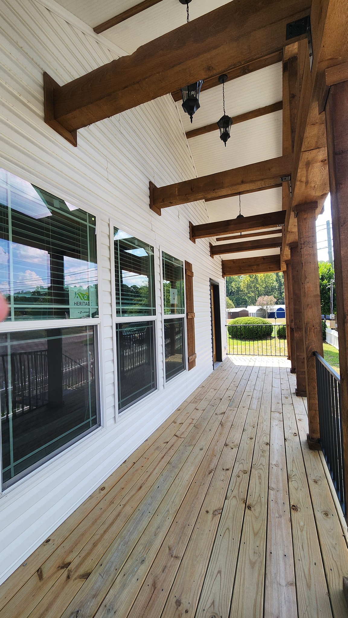 Covered wooden porch with white siding, large windows, and exposed beams. Sunlight filters in, casting shadows. Greenery visible in the background.