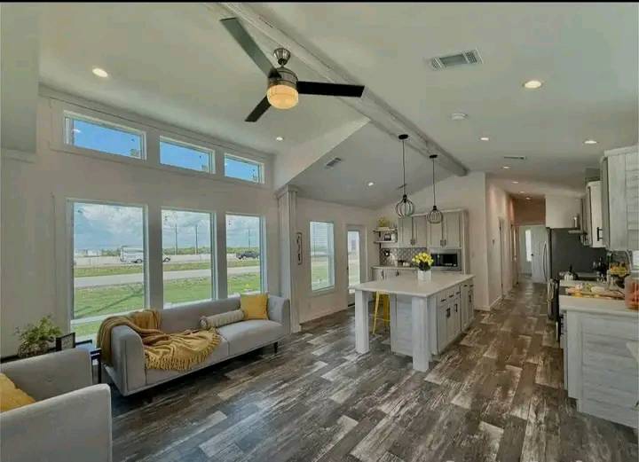 Bright, modern living area with large windows, a ceiling fan, gray couches, and a kitchen island. Sunlight enhances a warm, inviting atmosphere.