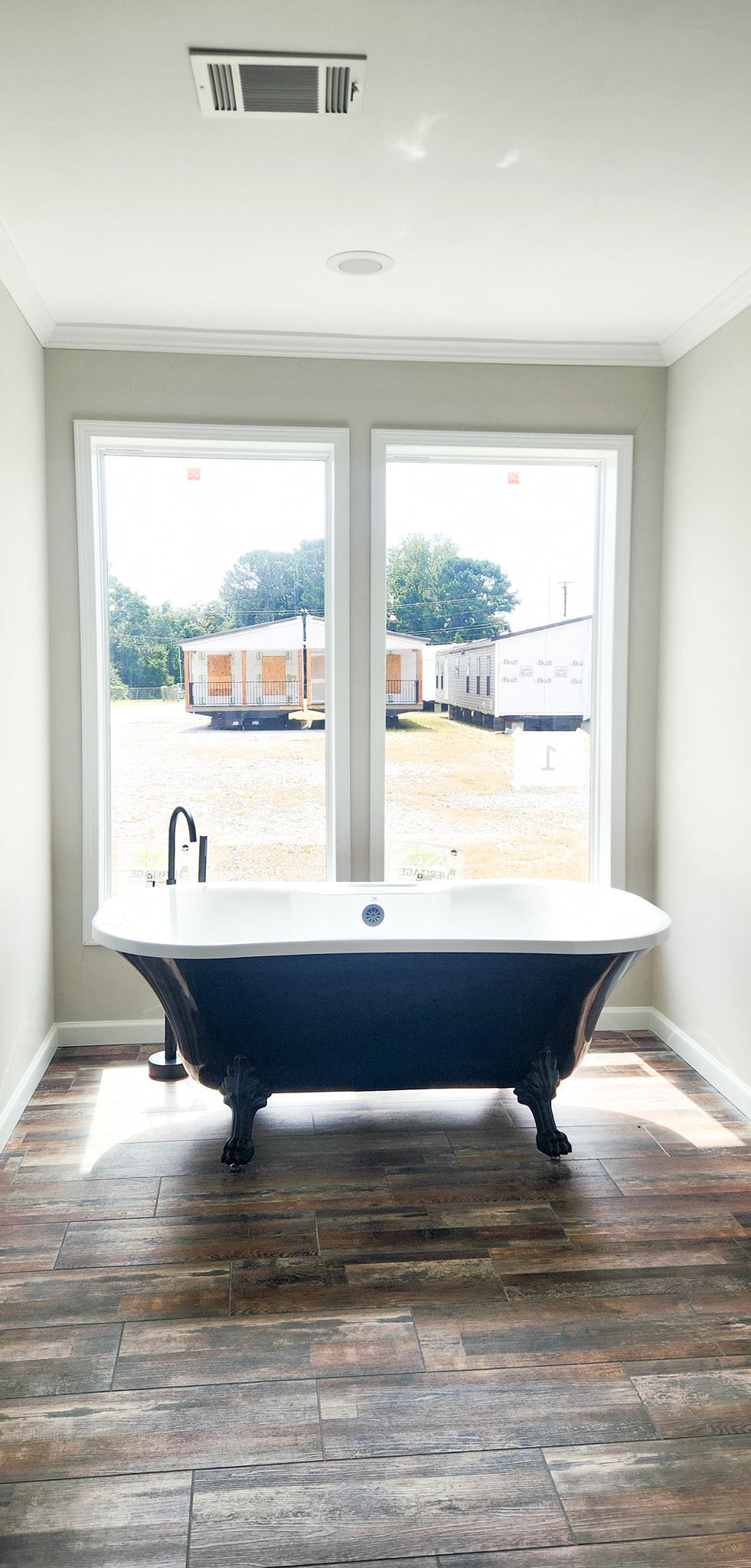 A serene bathroom features a vintage clawfoot bathtub set against two large windows. Sunlight streams in, illuminating the rustic wood floor and white walls, creating a tranquil and inviting atmosphere.