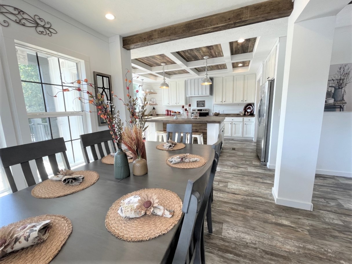 Modern farmhouse kitchen and dining area with wooden beams, white cabinets, and rustic decor. A dark table features woven placemats and floral napkins, exuding warmth.