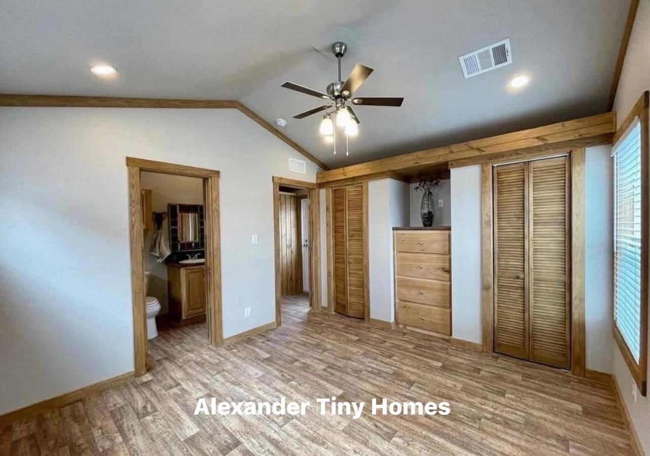 Spacious room with light wood paneling, a ceiling fan, and two open doorways leading to a bathroom and hallway. Text reads "Alexander Tiny Homes."