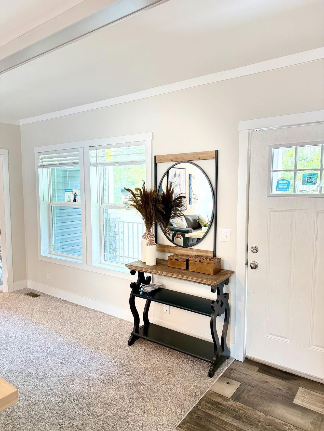 Entryway with a rustic console table, a round mirror above, and a decorative vase with dried plants. Natural light streams through adjacent windows.