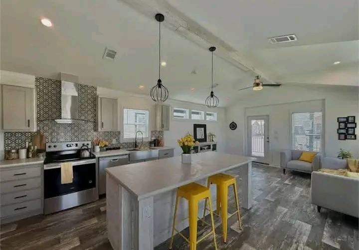 A modern kitchen and living area with light gray cabinetry, mosaic backsplash, and a stainless steel stove. A large island features yellow stools and pendant lights. A comfortable seating area with gray sofas and bright cushions is visible, creating a cozy and inviting atmosphere.