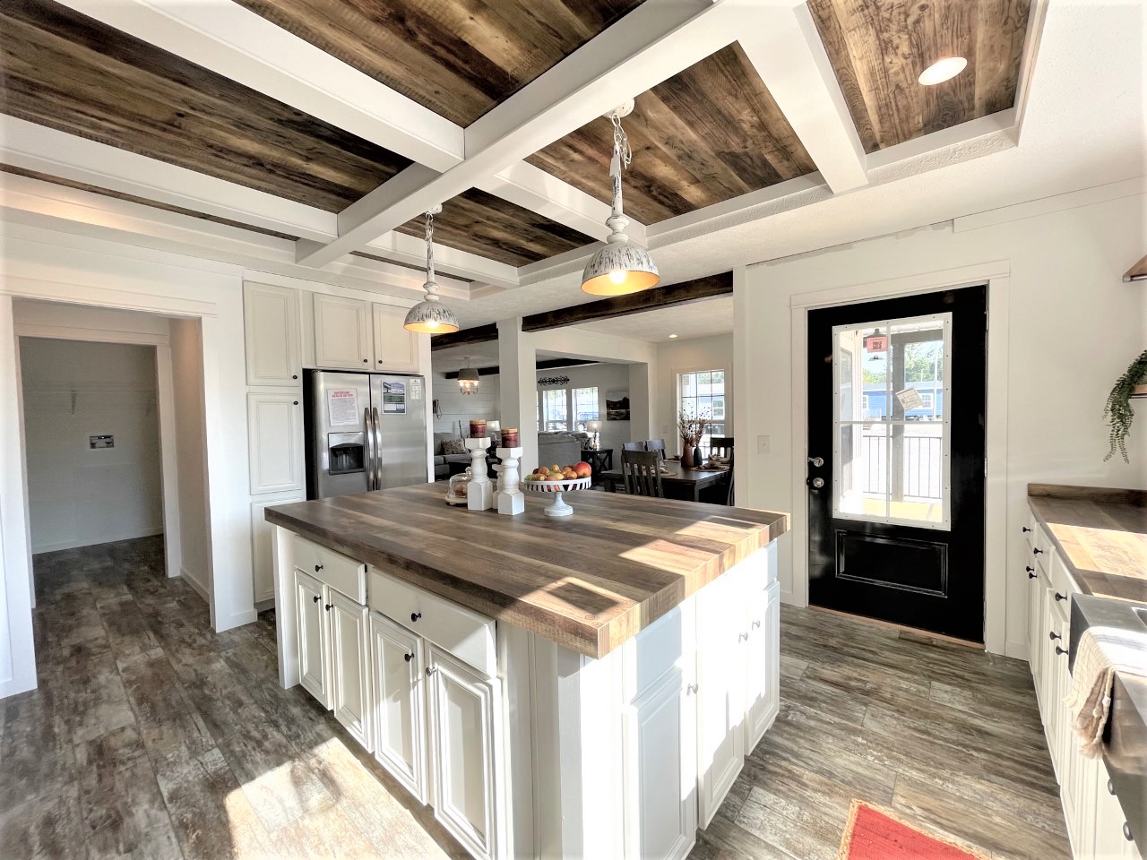 Bright kitchen with white cabinets, wooden countertops, and a rustic wooden ceiling. Hanging pendant lights add warmth. An open door reveals daylight.