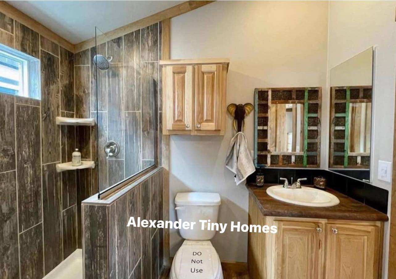 A compact, modern bathroom with wood accents. Features a shower with dark tiles, a wooden cabinet, and a sink with a mirror. Cozy, clean, and inviting.