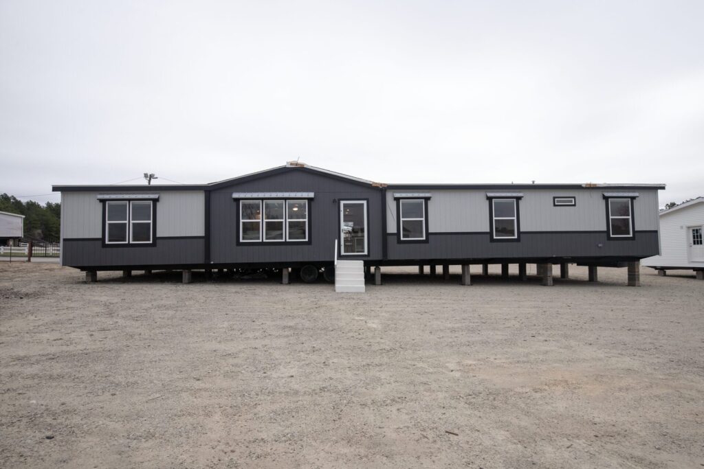 Modern gray manufactured home elevated on blocks, with multiple windows and small steps leading to the door, set on a gravel lot on a cloudy day.