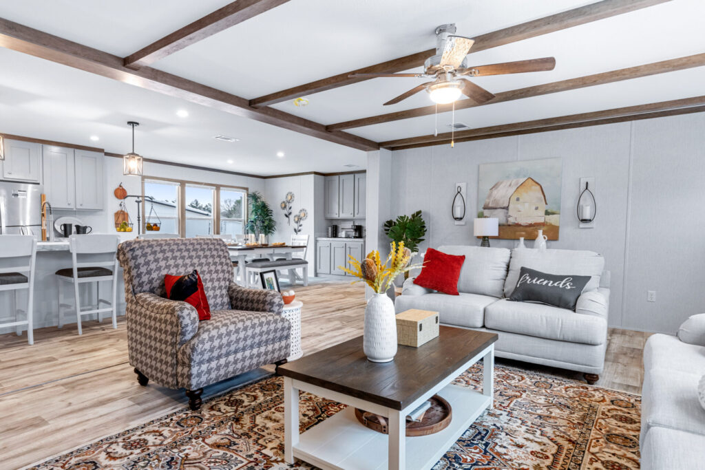 Spacious, cozy living room with gray sofas, red cushions, patterned rug, and a wooden coffee table. Ceiling fan above; kitchen visible in the background.