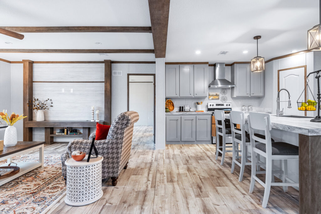 Modern kitchen with light gray cabinets, a central island with three chairs, pendant lights, stainless steel appliances, and wooden floors.