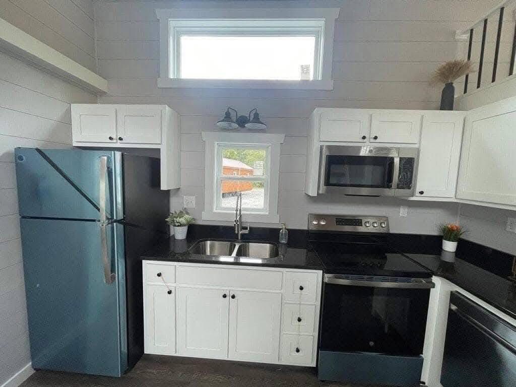 A modern kitchen with white cabinets, black countertops, a stainless steel fridge, microwave, and oven. Bright, with a window above the sink. Cozy and clean.