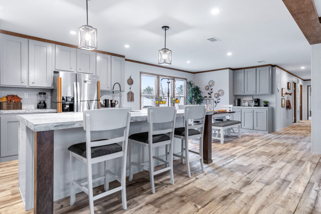 Spacious modern kitchen with light gray cabinets, marble countertops, and wooden floors. Pendant lights hang over a large island with white chairs. Cozy and inviting atmosphere.