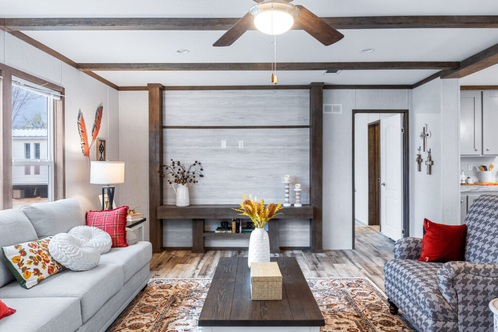 Modern living room with a gray sofa, colorful cushions, and a patterned armchair. A wooden coffee table holds a vase with yellow flowers. Ceiling fan above. Cozy and inviting atmosphere.
