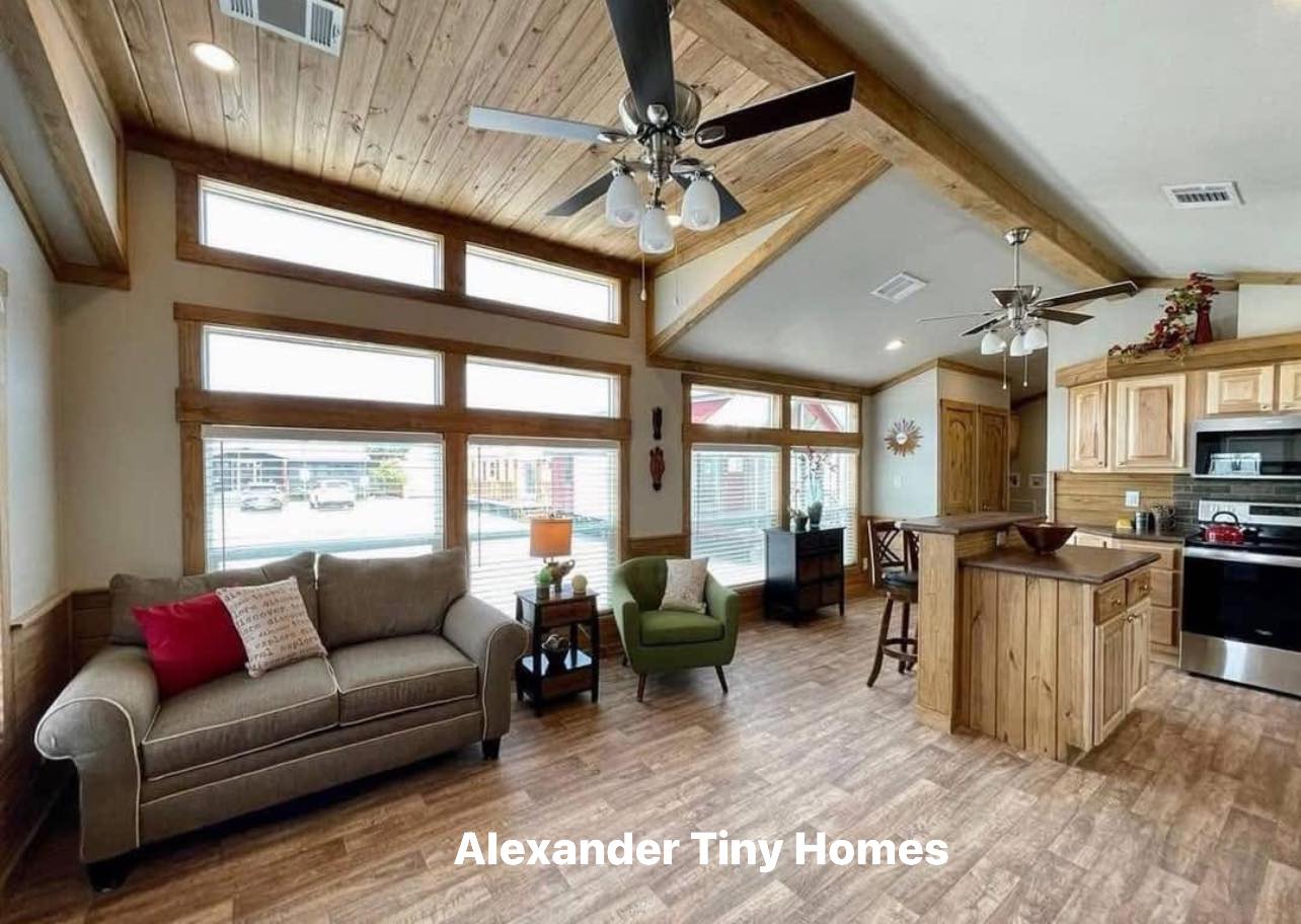 Spacious tiny home interior with wood accents, a cozy living area with a sofa and armchair, and a modern kitchen. Large windows fill the space with light.