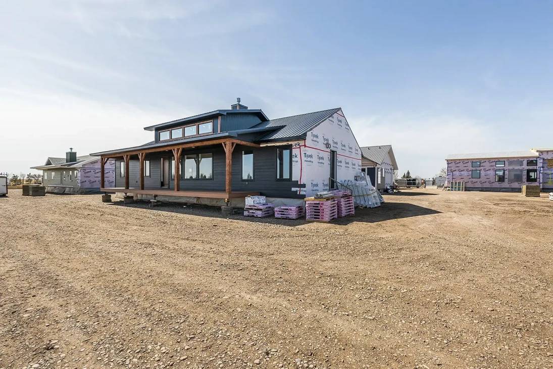 A modern house under construction stands on a dirt lot, with a wooden porch and low-pitched roof. The unfinished exterior is wrapped in sheathing materials.