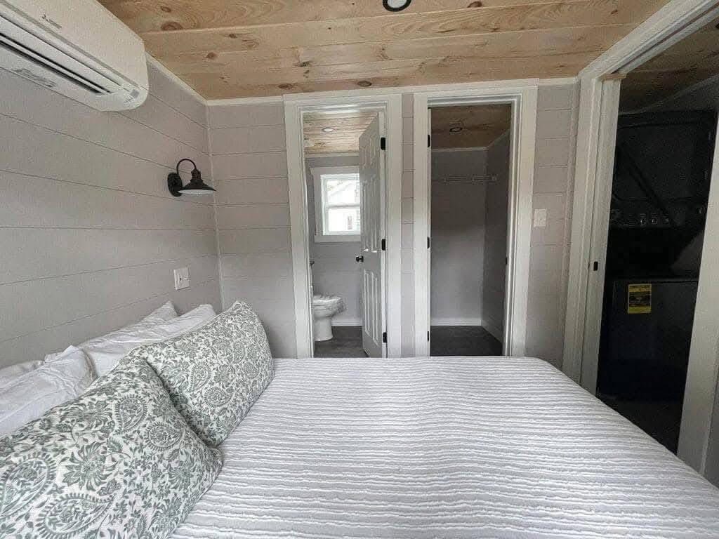 Cozy bedroom with a neatly made bed, floral pillows, light wood ceiling, two open doors leading to a bathroom and storage, and a wall-mounted AC.