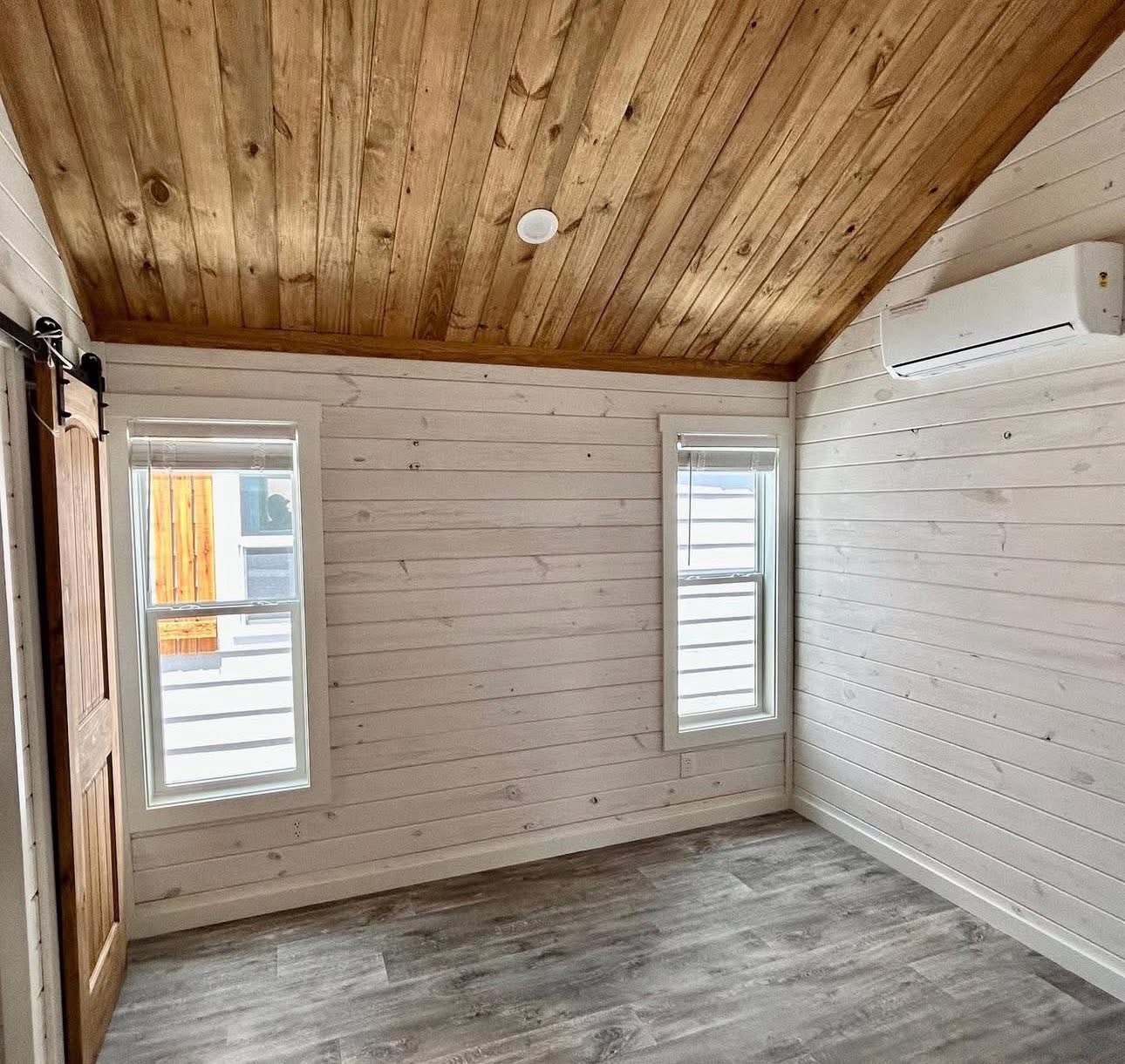Small empty room with light wood panel walls and ceiling. Two narrow windows with blinds, a wooden sliding door, and a wall-mounted air conditioner. Bright and minimalist.