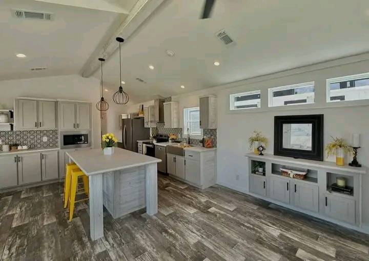 Modern kitchen with gray cabinets, an island with yellow stools, and pendant lights. Stylish decor on shelves, textured floor, and bright atmosphere.