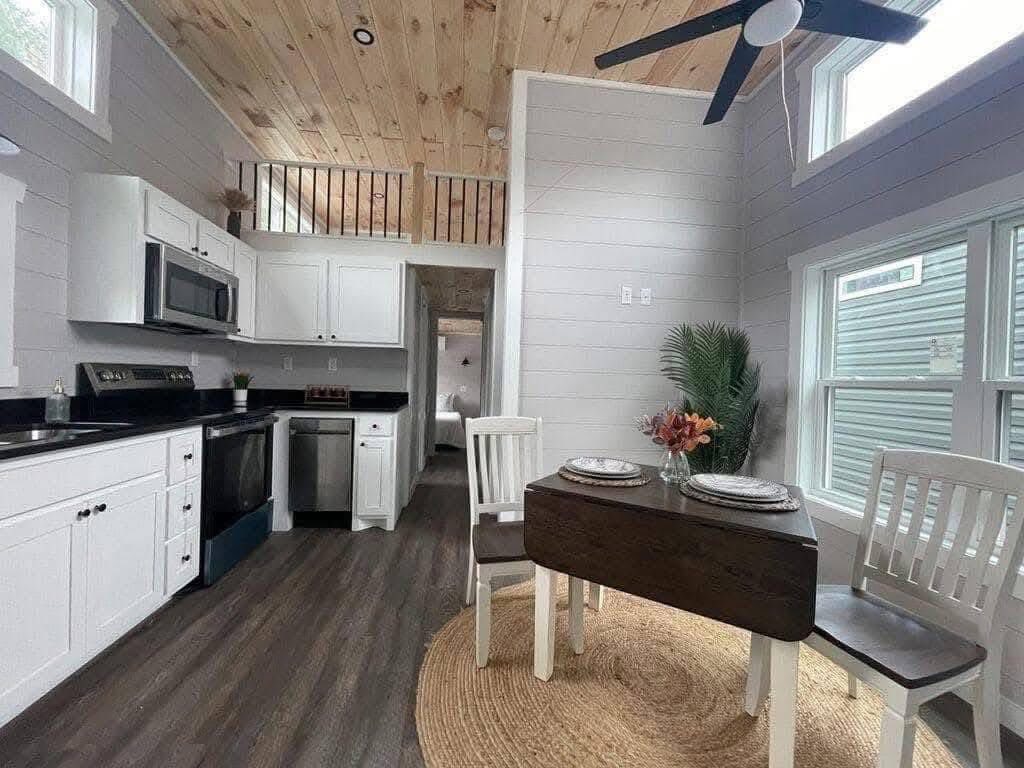 Modern tiny house interior with light wood ceiling and floors. White kitchen cabinets, black appliances, small dining table set, cozy, inviting atmosphere.