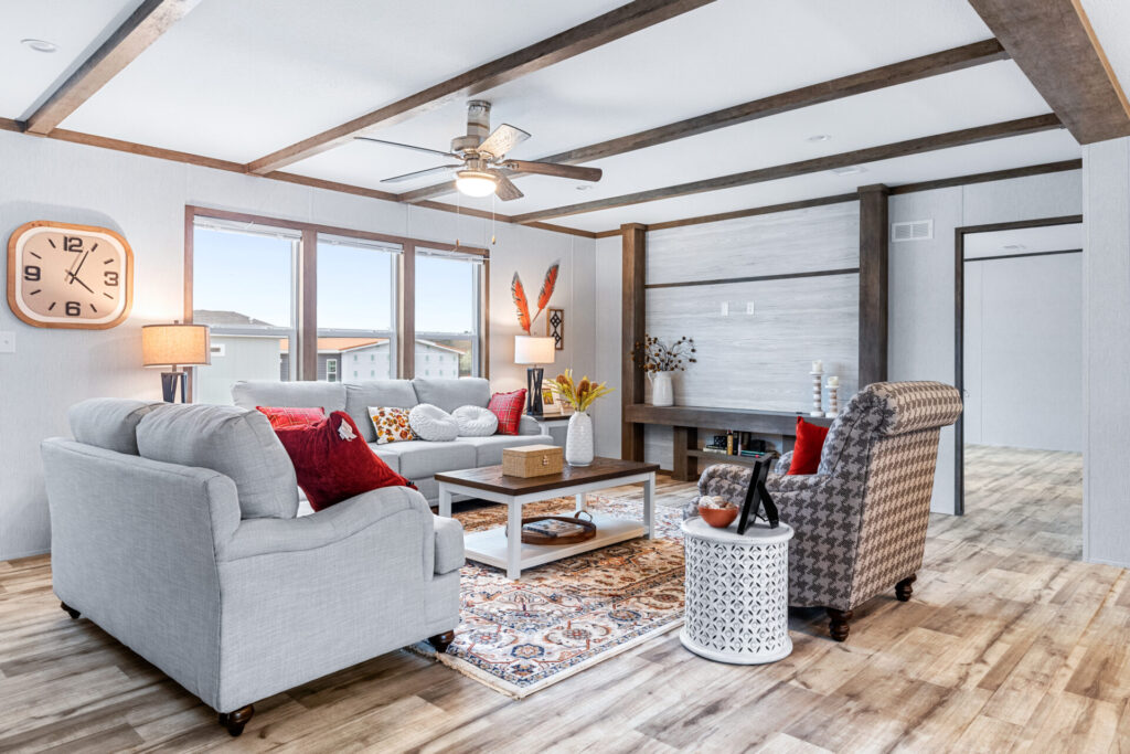 Stylish living room with gray sofa, patterned armchair, and colorful cushions. Wooden coffee table on a vibrant rug, large windows, and wall clock. Cozy ambiance.