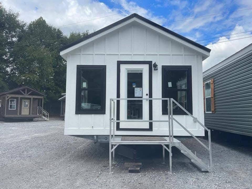 Small, white tiny house with black trim and a front porch. It has a door with large windows on either side. Overcast sky and trees in the background.