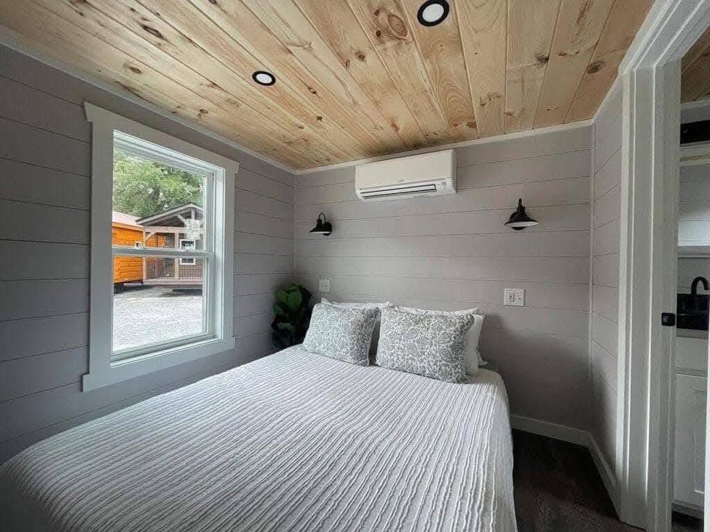 Cozy bedroom in a tiny house with light gray walls, a wooden ceiling, and a double bed with patterned pillows. A window shows a glimpse of an orange cabin outside.
