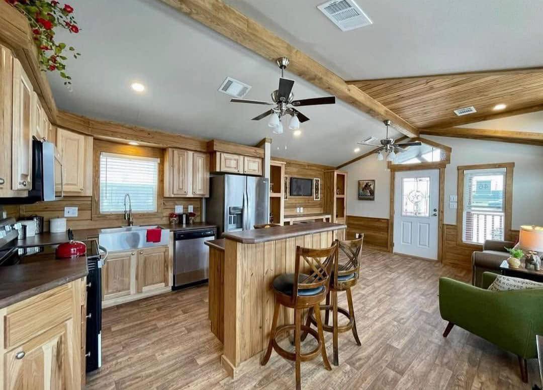 Spacious kitchen and living area with rustic wood finishes. Features stainless steel appliances, ceiling fans, bar stools, and cozy seating. Bright and welcoming.