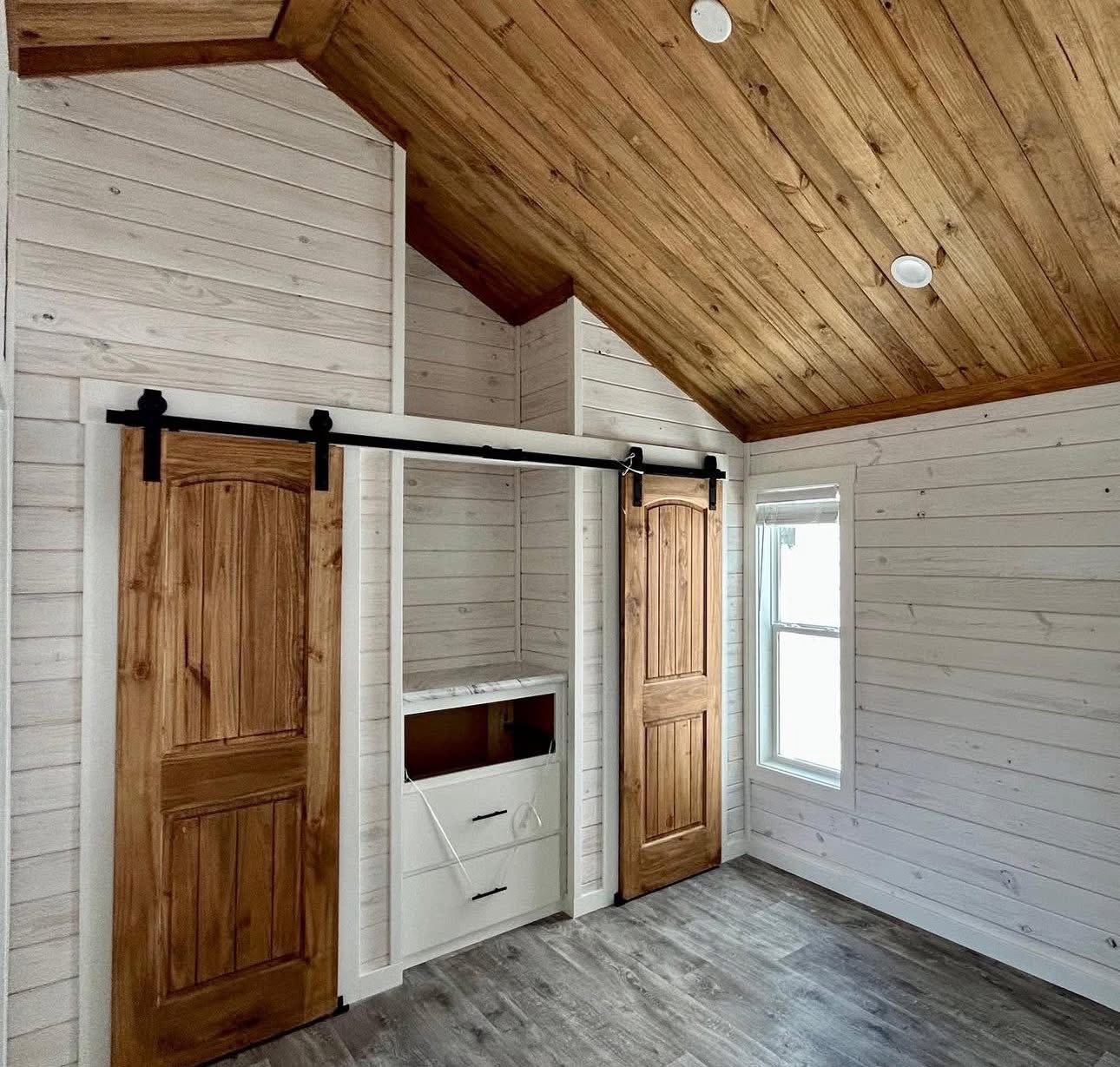 Cozy room with white wood panel walls and a sloped wooden ceiling. Features sliding barn doors, built-in cabinet, and a window with natural light.