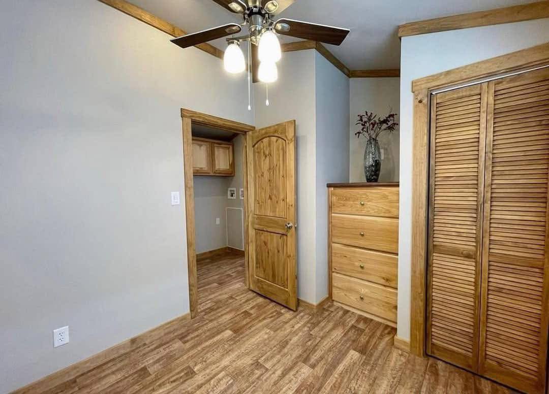 Cozy room with wood accents, featuring a ceiling fan, wooden door, dresser, closet, and light brown floor. Soft lighting creates a warm ambiance.