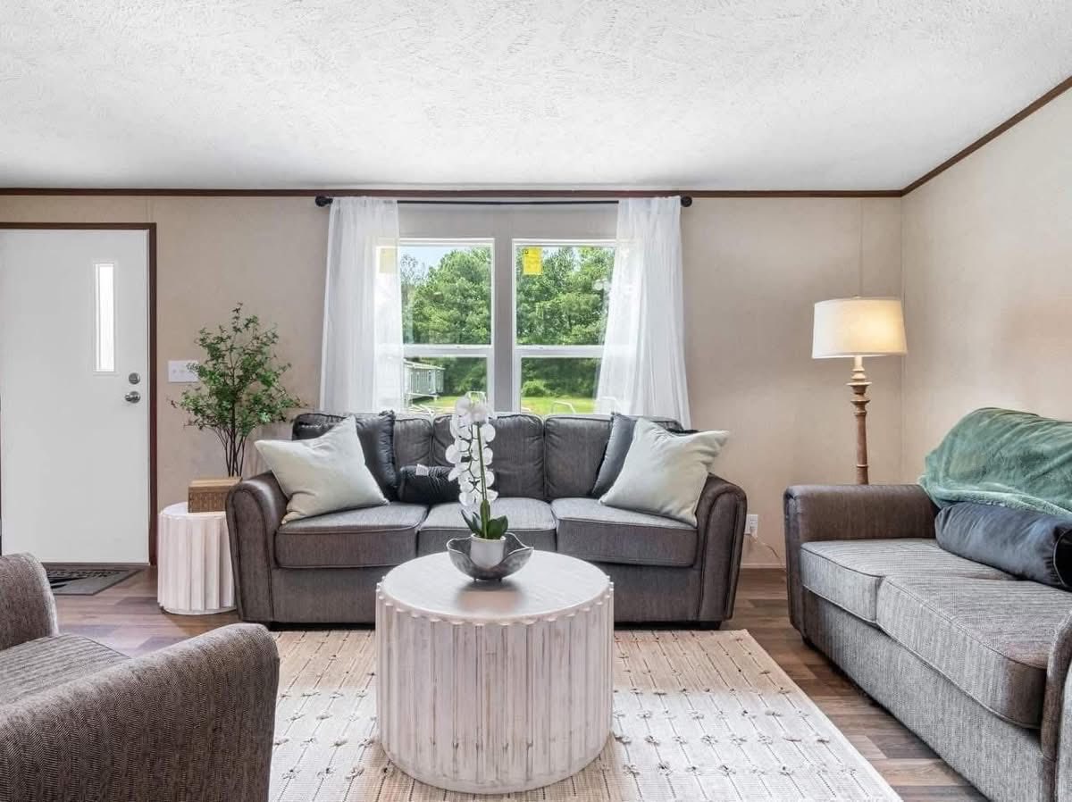 Modern living room with gray sofas, round white coffee table, orchid centerpiece, and floor lamp. Large window with sheer curtains and greenery outside.