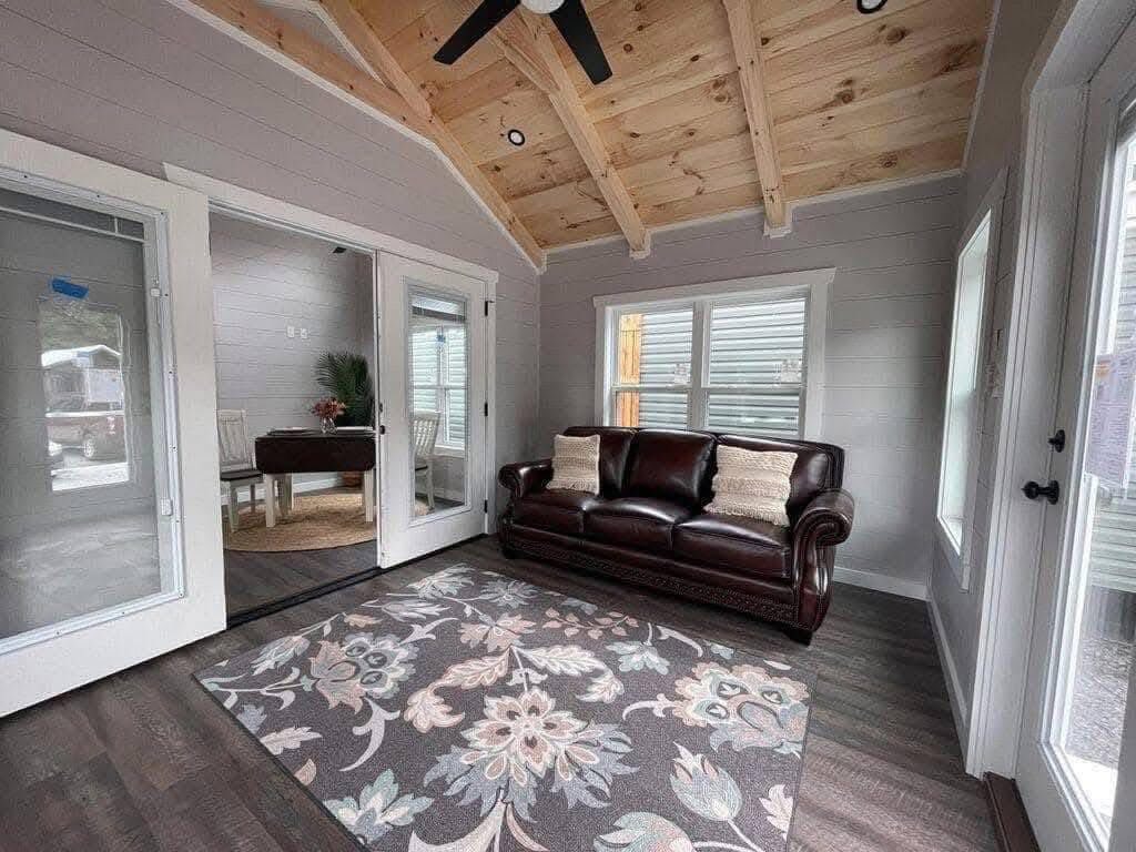 Cozy sunroom with wooden ceiling, brown leather couch, floral rug, and glass doors. Bright natural light creates a warm, inviting atmosphere.