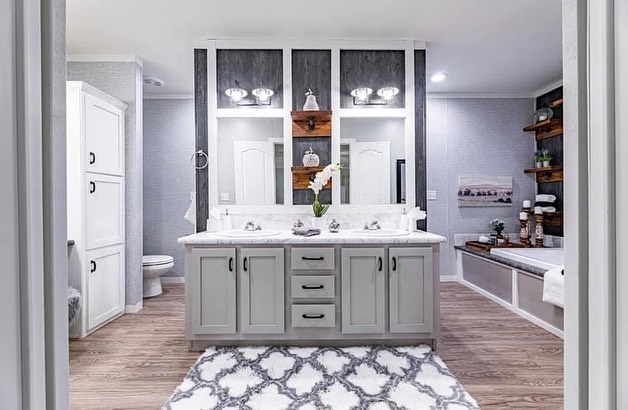 Bright bathroom with a dual-sink vanity and large mirrors, gray cabinets, wooden shelves, and soft lighting. Features light wood flooring and a patterned rug.