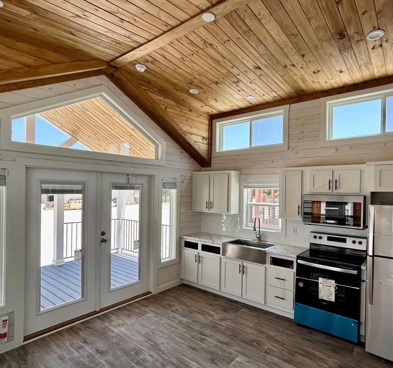 Modern kitchen interior with wooden vaulted ceiling, large triangular window, white cabinets, stainless steel appliances, and glass door to a deck. Cozy and bright.