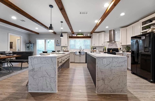Spacious modern kitchen with two marble islands, pendant lighting, and wooden beams. Features white cabinets, stainless steel appliances, and a large window.