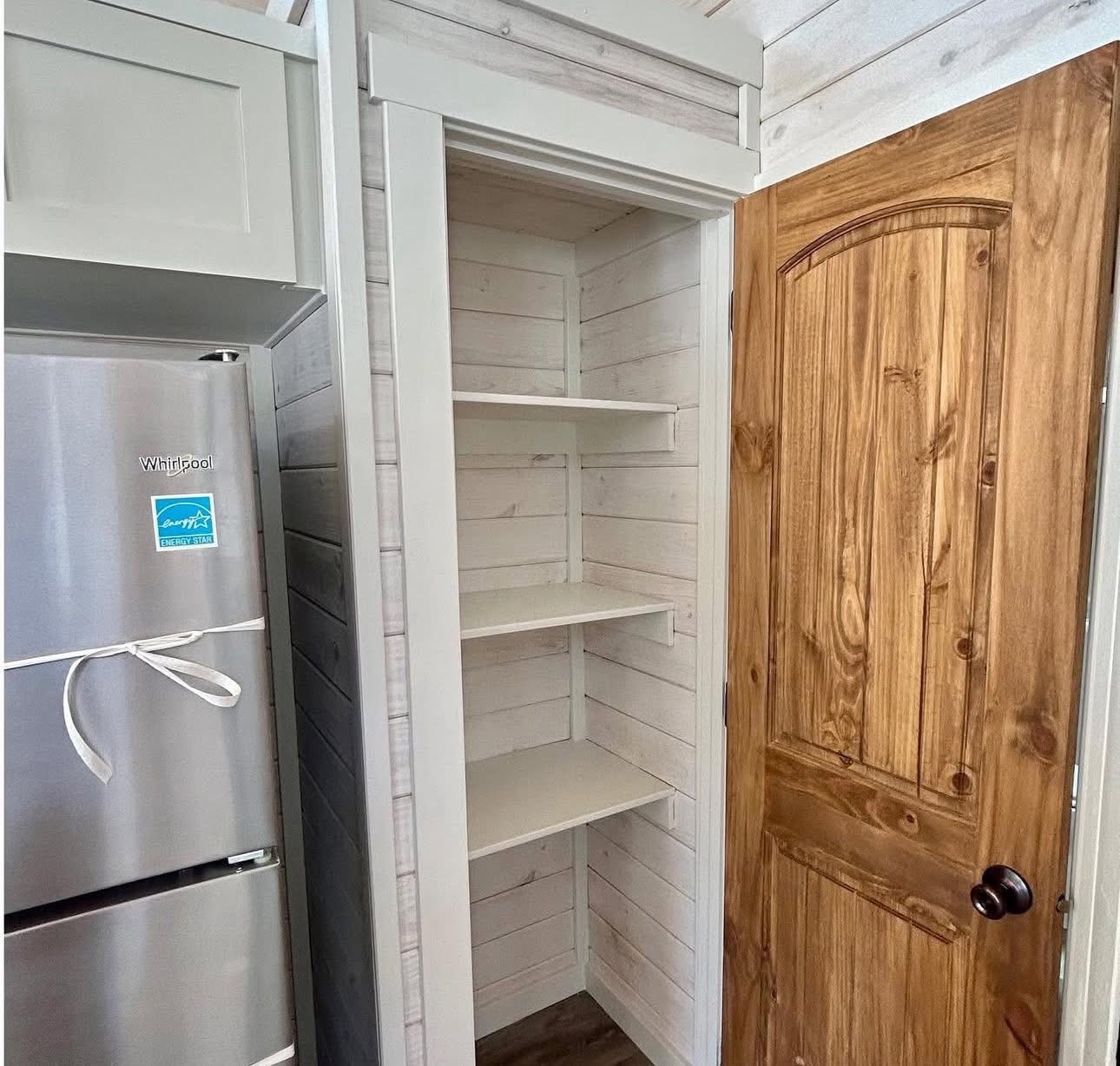 Compact kitchen pantry with white shiplap interior and three shelves beside a wooden door. Adjacent is a stainless steel refrigerator. Cozy and rustic.