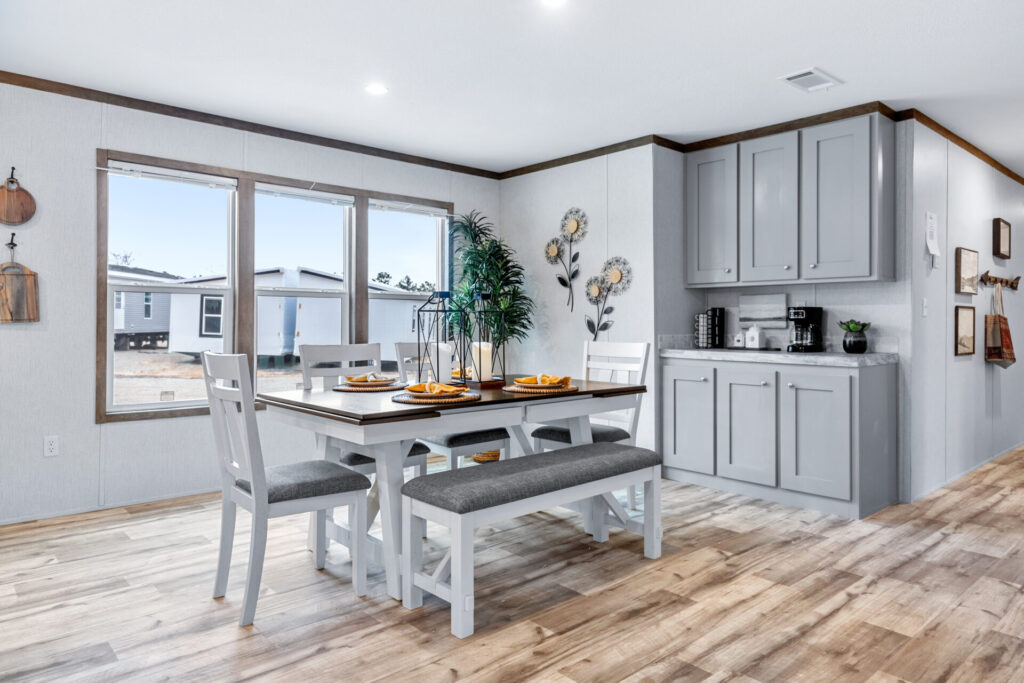 Bright dining area with large windows, a white table set for four with yellow accents, wooden floor, wall art, and a cabinet with kitchen appliances.