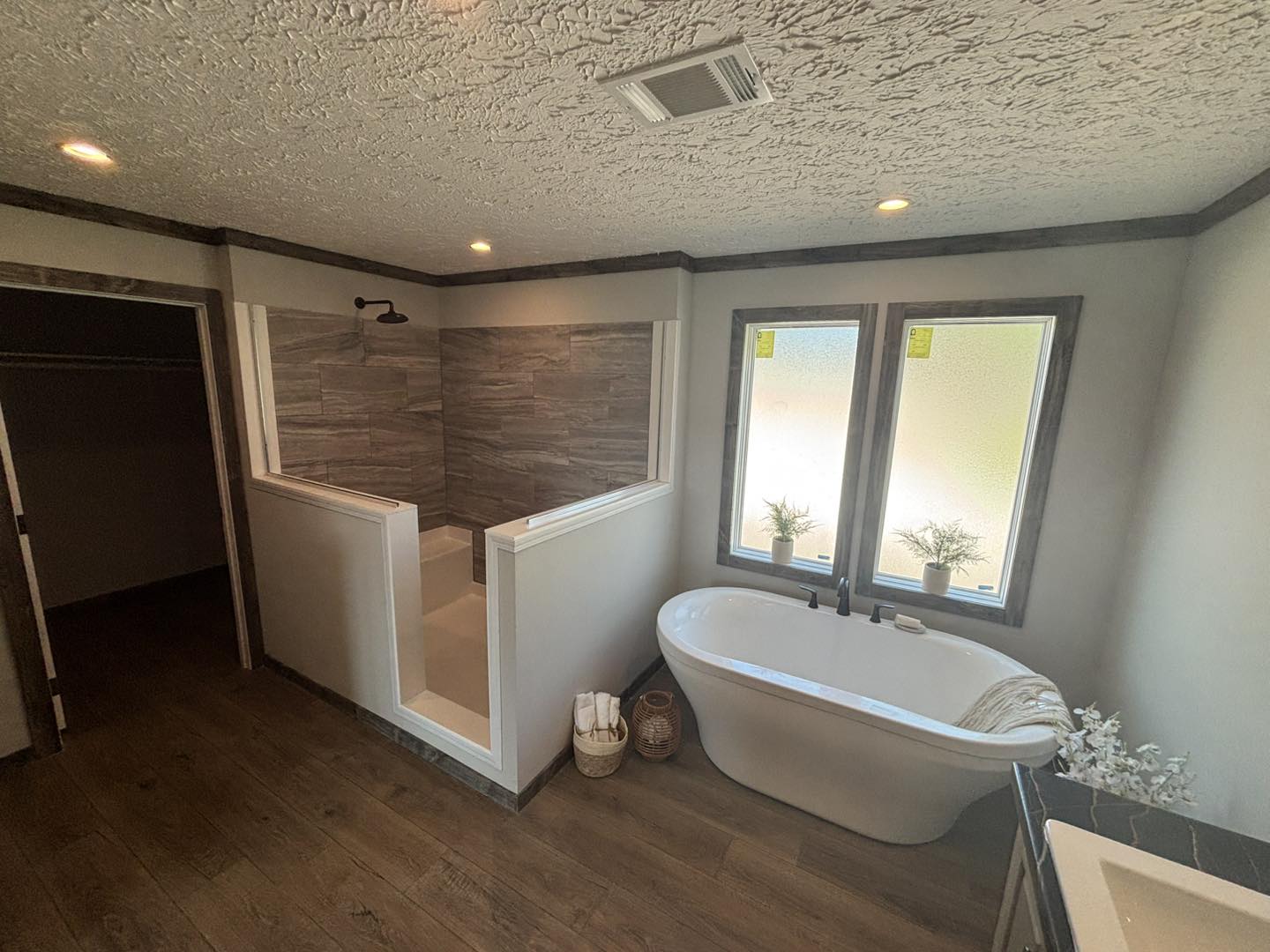 Spacious bathroom with a modern, gray-tiled open shower and a freestanding white bathtub. Large frosted windows and plant decor enhance the serene ambiance.