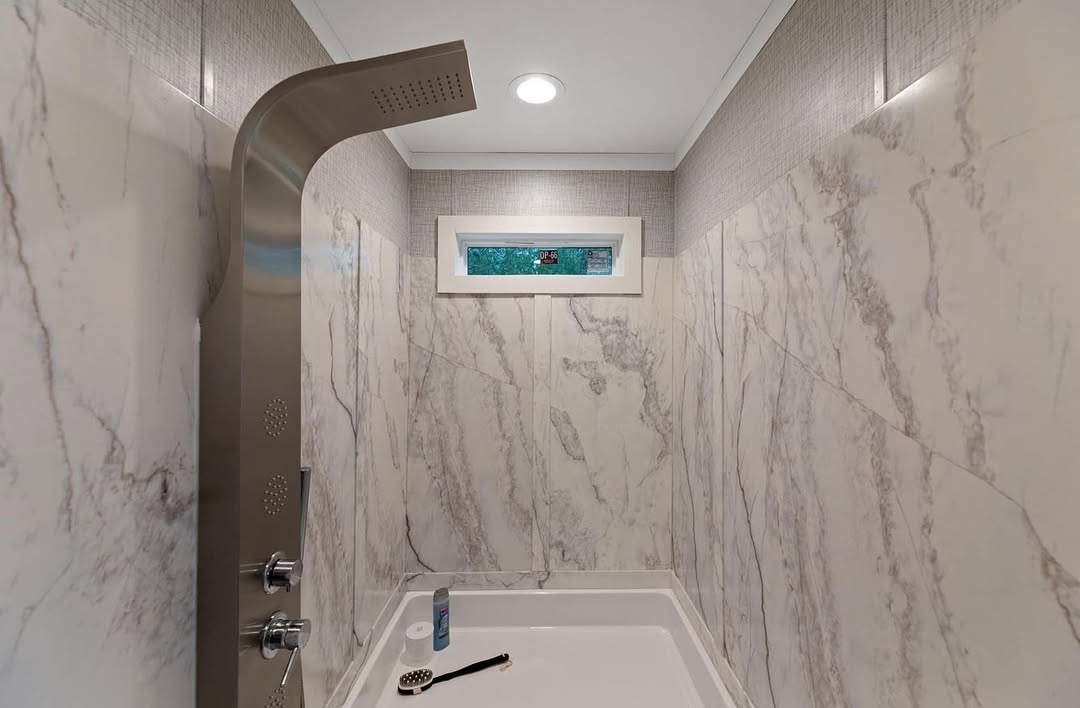 Modern shower with marble-patterned walls, silver rain shower panel, and a small window. The mood is clean and minimalistic. A brush and shampoo bottle rest on the white tub.